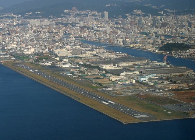 — — - Aerial view of Hiroshima-Nishi airport when it was still open to aircraft traffic. It has now been relegated as a heliport. Hiroshima city in the background.