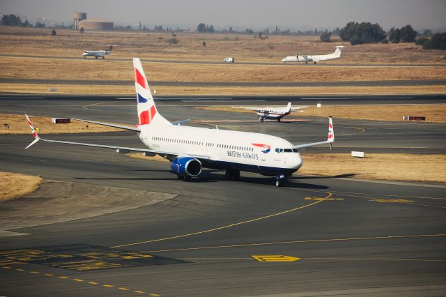 Boeing 737-800 (ZS-ZWG) - This Comair-Operated British Airways Boeing 737-8LD is about to turn off the taxiway to her parking bay after making the roughly 80 minute flight from Cape Town (FACT) to Johannesburg (FAOR) on Thursday, 1 August 2019. This picture was snapped at 11:43 in the morning, just on 15 minutes after touch-down on RWY 03R on a gloomy day. The sun popping through lit up her tail feathers quite nicely. *Click Full*