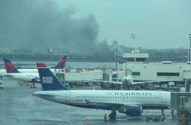 Airbus A319 (N749US) - US A-319 N749US prepares to push back at KLGA on April 1, 2011 as a large fire that ended up destroying a popular sports bar near Citifield, home of the New York Mets rages in the background less than 2 miles away..