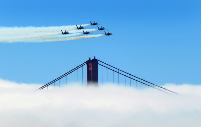 — — - Blue Angels in delta formation passing over the Golden Gate Bridge in the background.