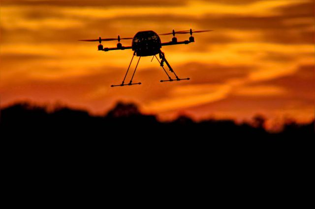 — — - A quadricopter drone hovers in the air as the sun sets.