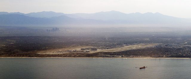 — — - The big picture of Los Angeles International Airport. View from rear view mirror, tugboats moving in on a jumbo oil tanker moored in the foreground, big buildings of downtown Los Angeles left center, looking north east from over the Pacific.