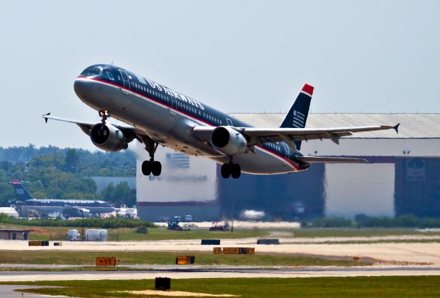 Airbus A321 (N181UW) - A hot afternoon takeoff from Runway 36 Center, Charlotte, North Carolina USA. You can see US Airways Charlotte Heavy Maintenance Hangar in the distance. It can easily hold an Airbus A330 in the center bay with room for six smaller planes if theyre parked creatively. A smaller "Line Hangar" (out of this photo but just to the left)is used by line maintenance for overnight routine inspections, engine changes, landing gear work and repairs to aircraft of an urgent nature. Specifically, if an aircraft is "broken" (in need of immediate maintenance) at the terminal, its brought over to the hangar by the "Supertug" (search N758US for Supertug photo). Highly trained and experienced aircraft maintenance technicians (AMTs) do what looks like miracles to get the airplane back on the gate for the flying public.