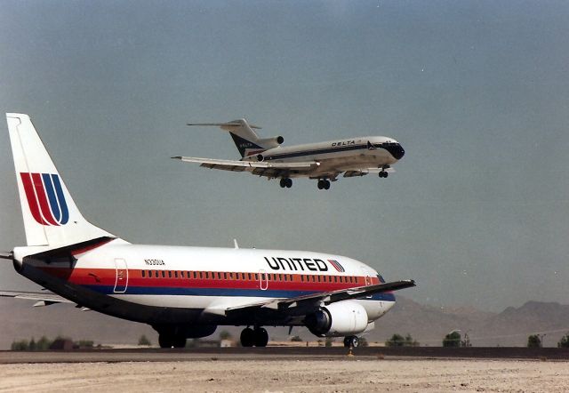 BOEING 737-300 (N330UA) - KLAS - Aug 1988 view, way before LAS made the airport improvements, one could park here north or south, of the 25 L R runways - complete with tumbleweed blowing around i the hot desert wind - and photograph airliners all day if you wished. Here a Delta 727 arrives 25 L as UAL 737 waits for its turn.