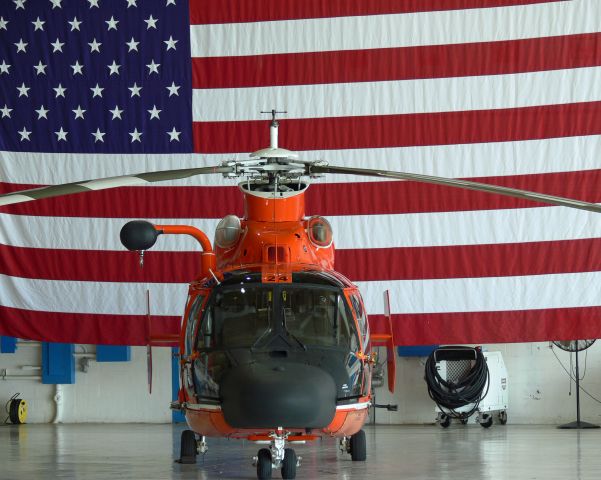 — — - HH-65 Dolphin inside the hangar with the flag as a background.