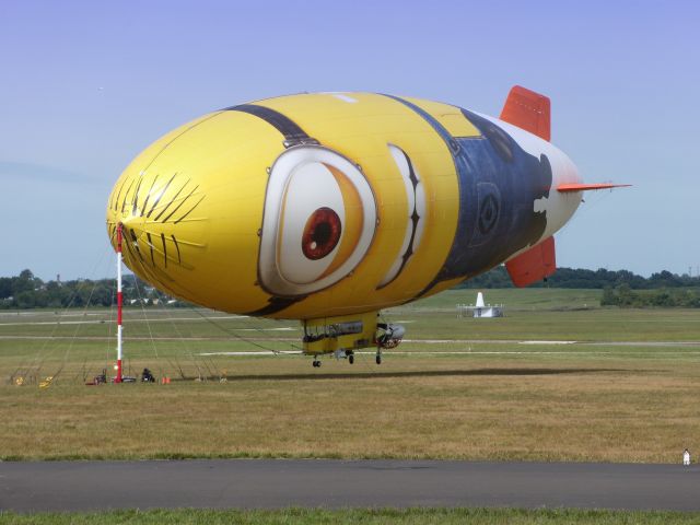 — — - This Airship image is representing a character from the Animated Movie Series "Despicable Me" and is called a "Minion".  The image is certainly strange enough to confuse the cat at the right corner of the picture. This Airship is tethered here in the Summer of 2013.