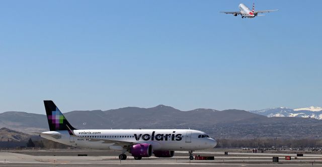 Airbus A320 (N533VL) - A Volaris Airbus (N533VL) on taxiway Charlie is at the 16L "Hold" line while that American flight in the background climbs away from 16L enroute to PHX.br /The VOI Bus was taxiing to the terminal after arriving from Guadalajara.