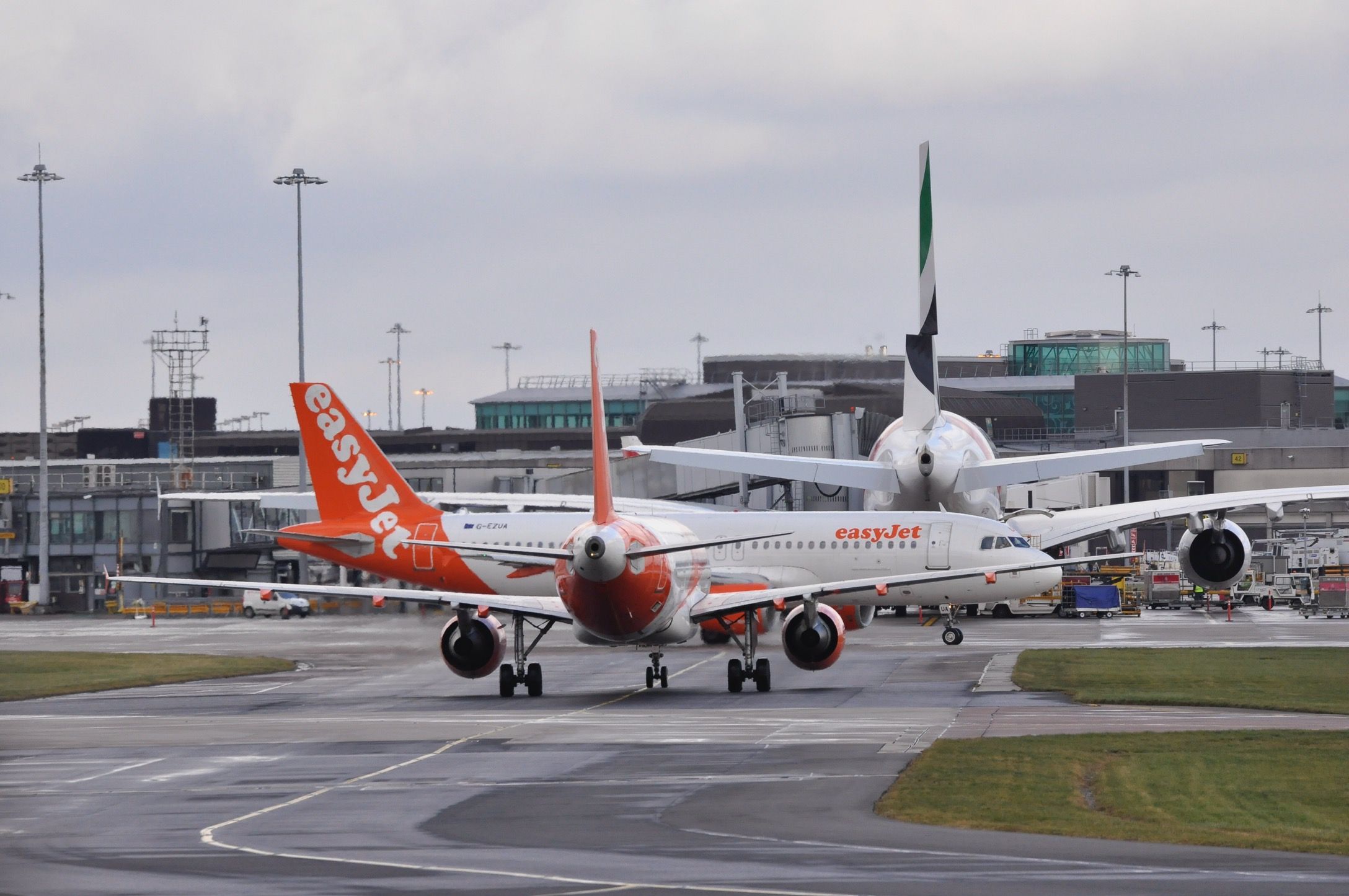 Airbus A320 (G-EZUA) - Retro liveried A320 taxis out of T1 terminal at Manchester as another Easy rolls towards it & an Emirates A380 waits to push back for DXB