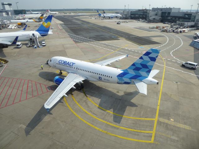 Airbus A319 (5B-DCU) - Cobalt Airways A319-100 5B-DCU standing at position T2 FRA. Photo made 27.08.2017 from off SkyLine train while on my way from T1 to T2.