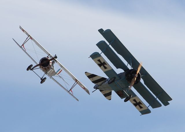 — — - A Tri Plane and Bi-Plane perform a mock dogfight at Duxford Air Museum.